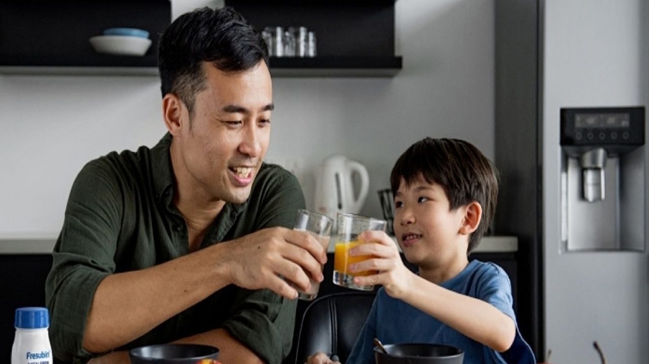 Adult and child raising glasses of orange-colored drinks at a kitchen table with bowls and fresh fruit.